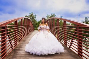 Quinceañera in a white tulle ball gown and tiara stands on a red-railed wooden footbridge under a partly cloudy sky.