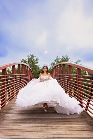 Quinceañera in a ruffled white ball gown and tiara walks across a red-railed wooden bridge under a cloudy blue sky.
