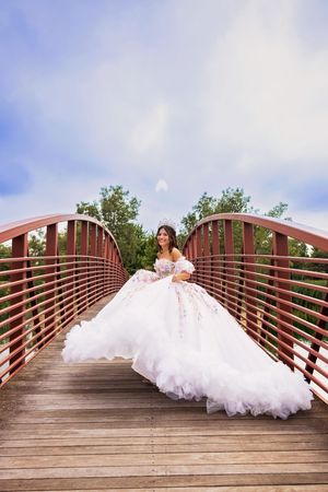 A quinceañera in a voluminous white floral ball gown sits and smiles on a wooden footbridge under a soft blue and cloudy sky.