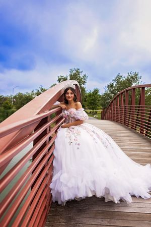 Young woman in a white off-the-shoulder floral ball gown posing on a red wooden footbridge beneath a blue sky.