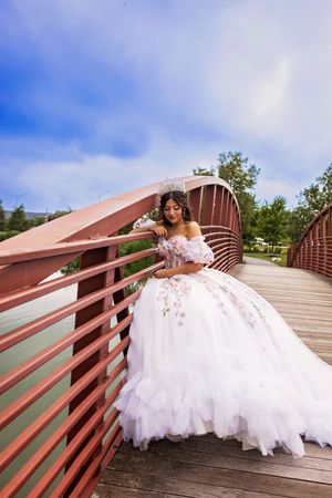 A young woman in a voluminous white tulle quinceañera gown with pink floral appliqués leans on a red bridge railing outdoors.