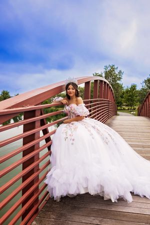 A quinceañera in a white ball gown with floral details and a tiara poses gracefully on a red wooden bridge beneath a blue sky.