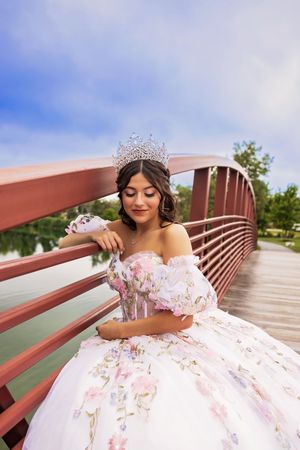 Close-up of a quinceañera wearing a sparkling tiara and an off-shoulder floral gown, resting her arm on a red bridge railing.