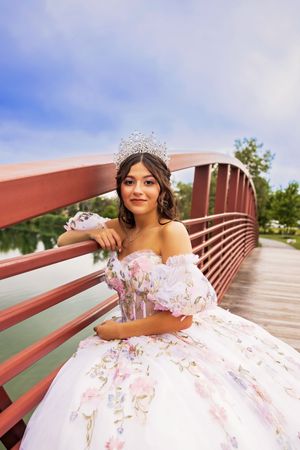 Quinceañera wearing a sparkling tiara and white gown with pink floral details poses on a red bridge by a lake.