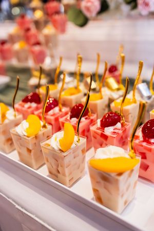 Rows of dessert cups filled with layered fruit jelly, topped with whipped cream, cherries and peach slices, served with gold spoons.