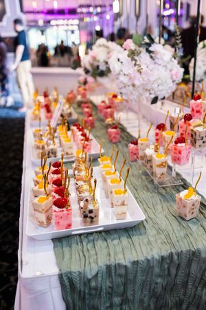 Elegant dessert buffet with layered fruit parfait cups topped with strawberries and mango on a sage green table runner near white floral cen