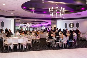 Elegant banquet hall with guests seated at round tables under a chandelier and purple ceiling lighting for a celebration.