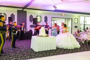 Quinceañera in a white ball gown stands beside a cake table as a mariachi band in uniform performs at the banquet hall reception.