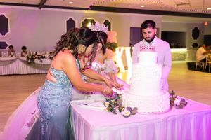 Quinceañera in a teal gown and tiara cuts a tiered white cake with a man in a light suit at a decorated reception hall.