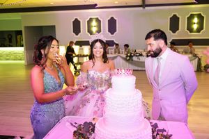 A quinceañera in a sparkling tiara and pink gown stands beside a tiered white cake with a young woman and a man in a pink suit.