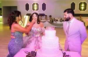 A woman in a sparkling dress feeds cake to a quinceañera beside a tiered white cake as a man in a pink suit looks on.