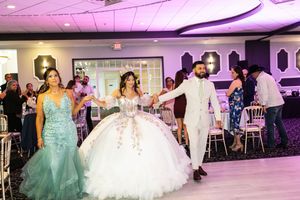 A quinceañera in a white ball gown dances hand in hand with a man in a white suit as guests watch at a reception hall.