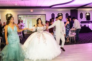 Quinceañera in a white beaded ball gown dances with a man in a light suit as a young woman in a teal gown watches.
