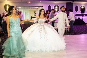 Quinceañera in a white ball gown holds hands with a woman in a teal gown and a man in a light suit at her reception.