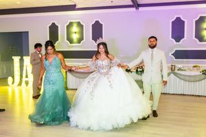 Quinceañera in a white ball gown and tiara holds hands with a woman in a teal dress and a man in a suit on a banquet hall dance floor.