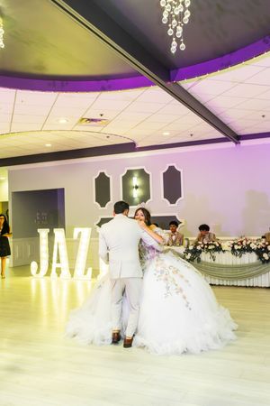 Quinceañera in a white floral ball gown dances with a man in a gray suit near glowing JAZI letters in a banquet hall.