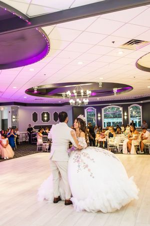 Quinceañera in a white embroidered ball gown and tiara dances with a partner on a banquet hall floor as guests watch.