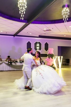 Quinceañera in a ruffled white ball gown dances with a man in a cream suit in a banquet hall with chandeliers and purple lighting.