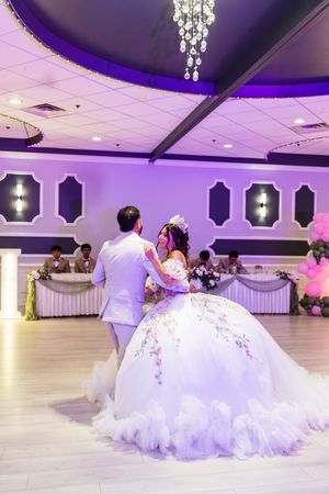 Quinceañera in a white floral ball gown dances with her partner in a purple banquet hall while guests watch from the head table.