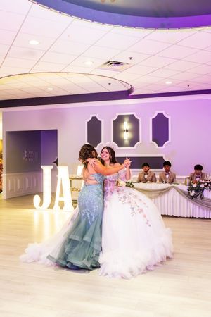 Quinceañera in a white embroidered ball gown embraces a young woman in a teal gown on the dance floor beside a lit letter display.
