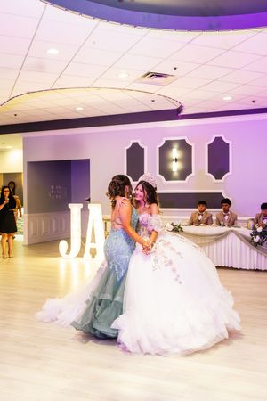 Two young women in a teal gown and a white quinceañera dress share an emotional embrace at a purple-lit reception hall.