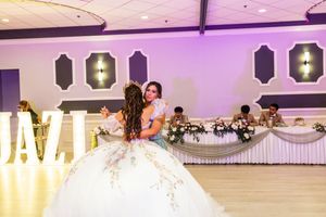 Quinceañera in a white floral gown embraces a guest on the dance floor of a banquet hall with a decorated head table behind them.