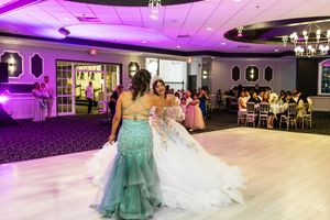 Two women in elegant gowns, one teal and one white ballgown, dance together on a ballroom floor as guests watch from tables.