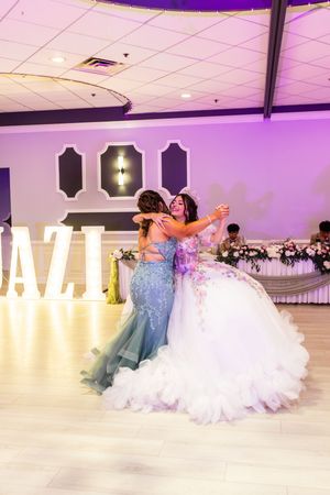 Quinceañera in a white ruffled ball gown embraces a woman in a teal beaded dress on the dance floor at a purple-lit reception hall.