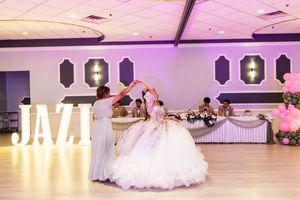 Quinceañera in a voluminous white gown dances with a family member in a banquet hall with a lit JAZ sign and decorated head table.