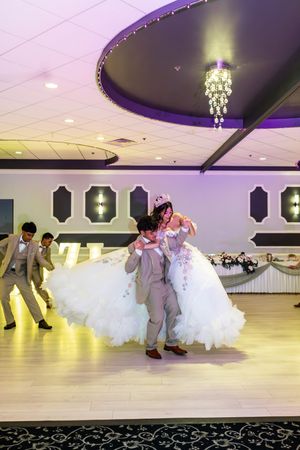 Quinceañera in a tiara and white tulle gown is lifted by a young man in a tan suit during a choreographed dance at her celebration.