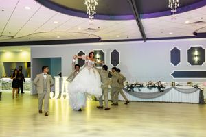 Quinceañera in a white ball gown is lifted and twirled by escorts in tan suits on the dance floor of a decorated banquet hall.