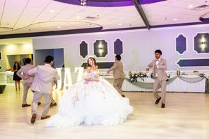 Quinceañera in a sparkling white ball gown dances a choreographed routine with chambelanes in tan suits on the banquet hall dance floor.