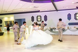 Quinceañera in a voluminous white ball gown dances with her court of chambelanes in tan suits in an elegant banquet hall.