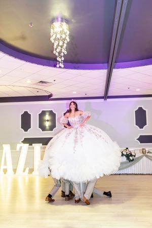 Quinceañera in a voluminous white gown is lifted high by her chambelanes during a choreographed dance beneath a crystal chandelier.