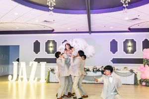 Groomsmen in light gray suits lift a young woman in a white dress above the dance floor beside a glowing JAZZ sign at a quinceañera.