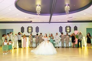 Quinceañera in a white ball gown stands on a polished dance floor surrounded by family and court members in a elegant banquet hall.