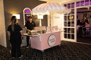 Pink dessert cart with a striped umbrella and treats on display, with a vendor in black standing at a party venue.