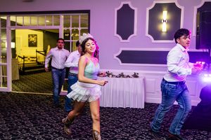 Young woman in a light blue dress and tiara dances at a quinceañera party while guests in jeans and dress shirts watch nearby.