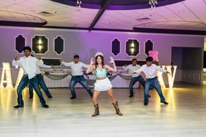 Quinceañera in a white skirt and cowboy boots performs a choreographed dance with male dancers on a reception dance floor.
