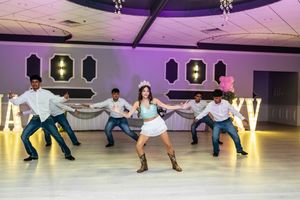 Quinceañera in a tiara and cowboy boots performs a choreographed dance with a group of chambelanes on a reception hall dance floor.