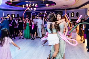 Two girls in formal gowns embrace on a banquet hall dance floor while guests dance around them with pink balloon twists.