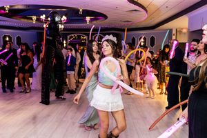 Quinceañera in a white skirt and tiara dances with guests wearing glow accessories on the dance floor at her celebration.