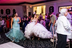 Quinceañera in a ruffled white gown walks across a reception hall beside a woman in a teal mermaid dress as guests look on.