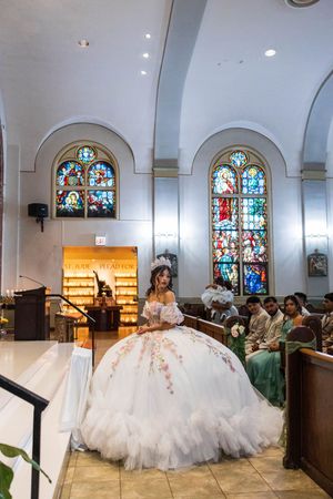 Quinceañera in a floral tulle ball gown and tiara stands in a church with stained glass windows as guests sit in the pews.