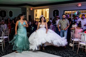 Quinceañera in a white ruffled ball gown walks across the dance floor as guests and a woman in a teal gown look on.