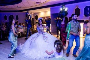 Quinceañera in a sparkling white embroidered ball gown dances with guests while a photographer captures the lively celebration.