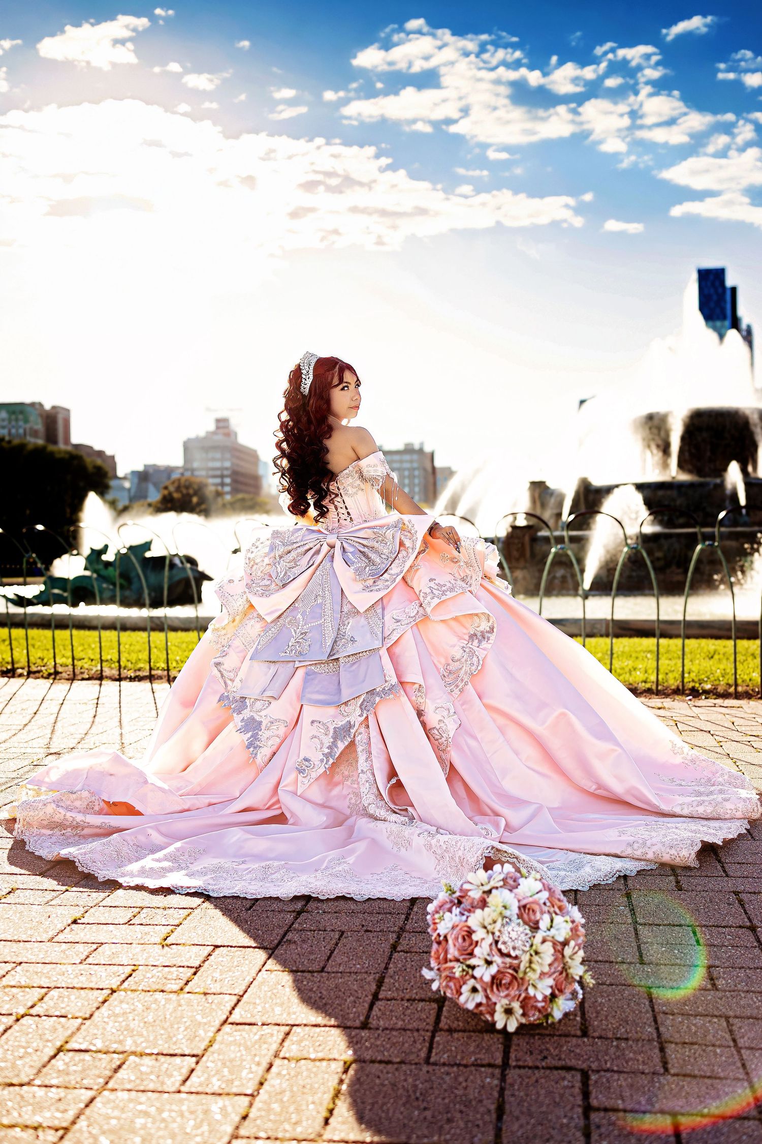 Woman in a pink princess ball gown with a large bow looks back beside sunlit park fountains, a bouquet of roses on the brick pavement.