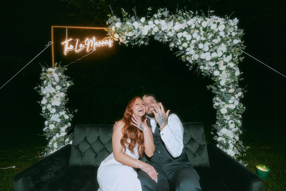 Newlywed couple shares intimate moment under illuminated floral arch with neon sign at night.