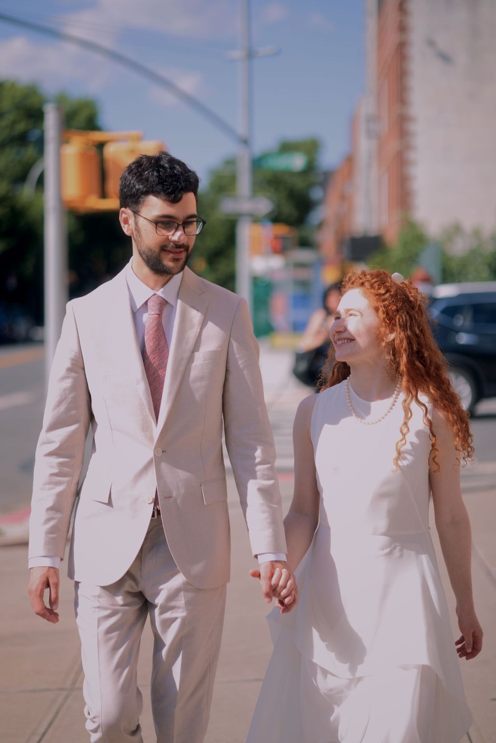 Couple in matching light-colored formal wear walking together on sunny city sidewalk.