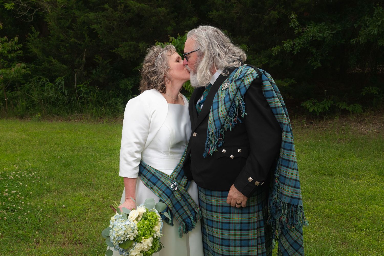 Couple in traditional Scottish wedding attire sharing a kiss in a green meadow.